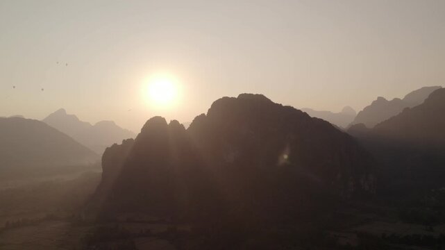 Aerial panning view of the setting Sun behind large mountains with tropical landscape and paragliders in the background.