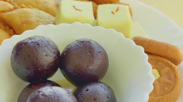 Turntable closeup focusing on glossy, syrup-soaked gulab jamun in a bowl, with barfi, peda, and gujiya highlighting rich shine, smooth texture, and indulgent sweetness.