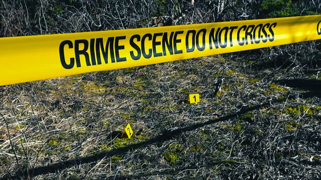 Yellow crime scene do not cross tape restricts access to an outdoor area where numbered evidence markers indicate the location of found clues, possibly bullets or shell casings, on the ground