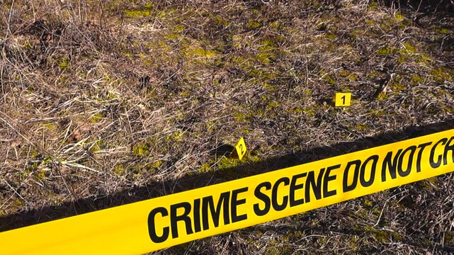 Yellow police tape with the words Crime Scene Do Not Cross cordoning off an outdoor area with numbered evidence markers placed on the dry grass, indicating a forensic investigation