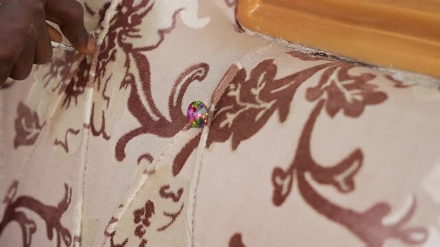 Upholstery repair closeup showing hands securing decorative tack into patterned fabric, upholsterer attaching trim to wooden frame, bead and stud embellishment, careful handwork with needle