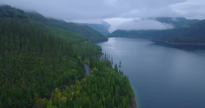 Misty mountain lake bordered by dense evergreen forest with low clouds drifting across the hills and calm reflective water, aerial drone peaceful background