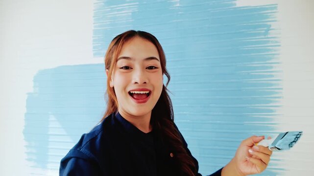 Smiling woman holding a paintbrush in front of a wall being painted light blue, mid-DIY home makeover