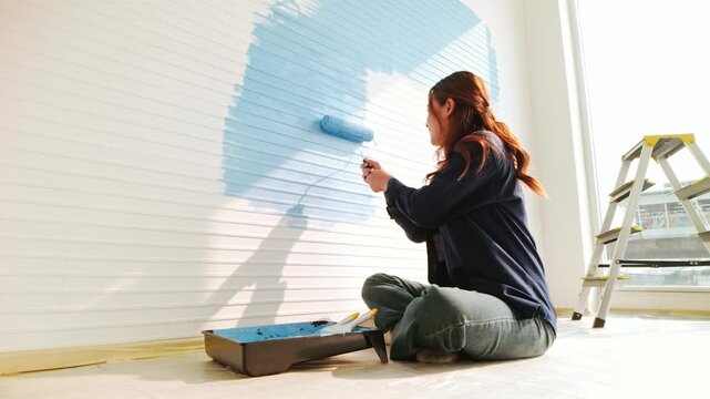 A woman sits on the floor using a roller to paint a white paneled wall light blue during a bright home DIY project, with a ladder and paint tray on a drop cloth nearby