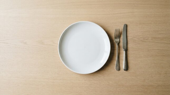 A white plate sits on a wooden table beside a fork and knife. The table surface is smooth and light in color. The setting is simple and ready for food