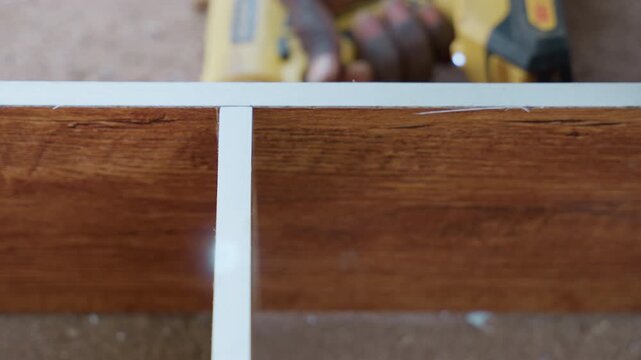 Closeup trim installation on stained wood, white edging aligned in groove with steady hands, drill resting in background, surface grain and clean lines visible, precise finish for cabinetry