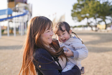 Fototapeta premium Mother hugs daughter warmly at park. Sunlight glints through her long, golden hair. Child beams, arms wrapped tight around her. Soft laughter echoes through the quiet playground