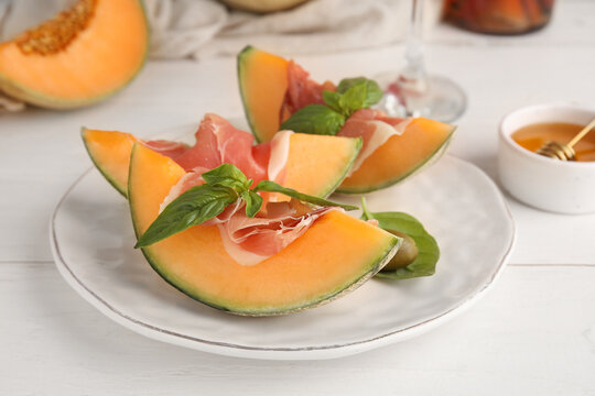 Plate of fresh melon pieces with delicious prosciutto and basil leaves on white wooden background, closeup