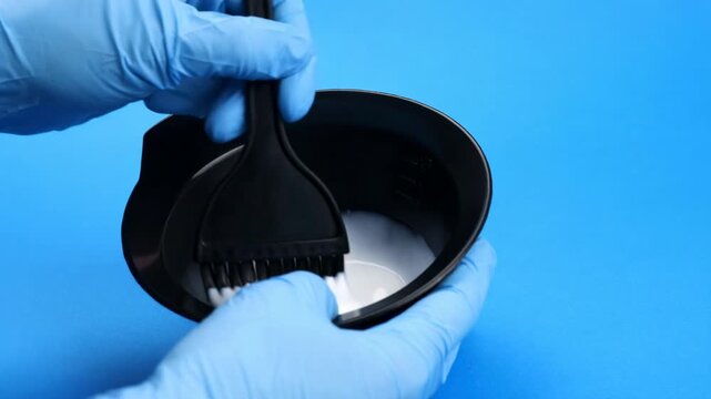 Professional hairdresser with gloves mixing hair dye in a bowl with a brush against a blue background