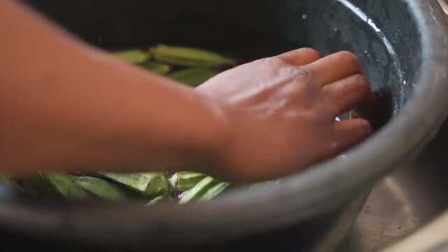 Wet hands washing green okra in black basin, closeup of fingers rinsing pods under running water, soapy bubbles and gentle motion, domestic kitchen prep and tactile routine before cooking