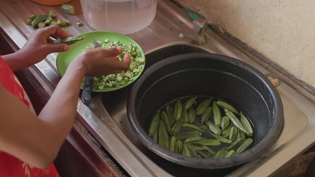 Black woman shelling peas at sink, transferring fresh peas into green bowl while empty pods collect in dark pot, warm natural light, closeup on careful hands, slow domestic routine, textured counter