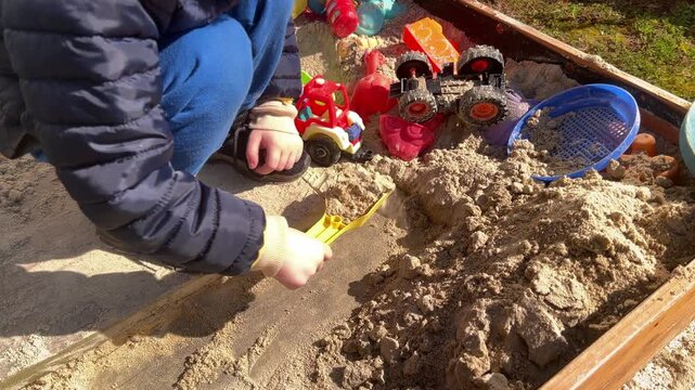 Toddler using yellow shovel to push red tractor before digging sand in wooden sandbox