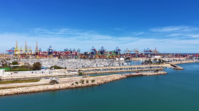 Elevated view of Valencia's large container terminal with colorful stacked cargo and cranes beside a sailboat marina on the Mediterranean coast