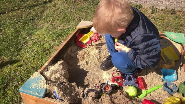 Little boy using yellow shovel to move sand in wooden sandbox surrounded by colorful toys
