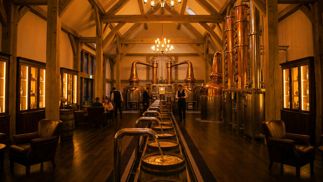 Inside a beautifully illuminated distillery showcasing massive copper pot stills and spirit filling into wooden barrels with staff present.