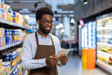 African american male employee wearing an apron and eyeglasses standing inside a grocery store aisle, using a digital tablet to manage inventory and check stock, smiling as he works © Liubomir