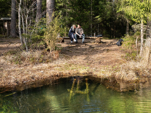 A young couple sitting on a wooden bench by the crystal clear turquoise Antu lake in Estonia forest during springtime.