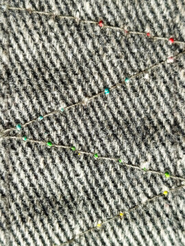 Detalle de hilos con peque&ntilde;as cuentas de colores dispuestas en zigzag sobre una base de tela gris con textura rayada
