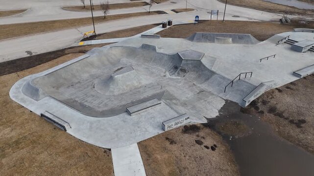 Drone orbiting right around a newly constructed concrete skate park, showing rails, ramps, brown spring grass, and water in a nearby ditch under clear skies.