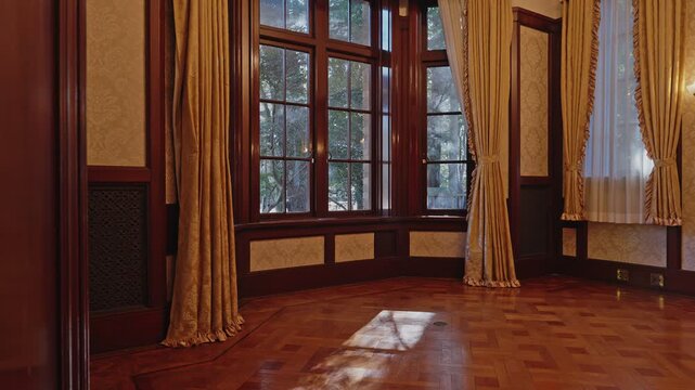 Interior view of a grand historic room featuring large wooden windows, golden curtains, and sunlight reflecting on the polished parquet floor.