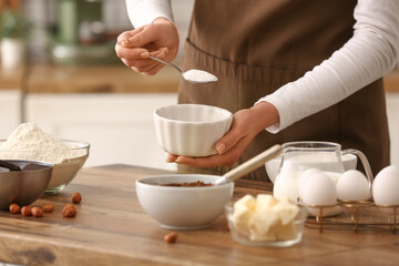 Fototapeta premium Woman adding sugar into bowl for preparing chocolate brownie at table in kitchen. Closeup