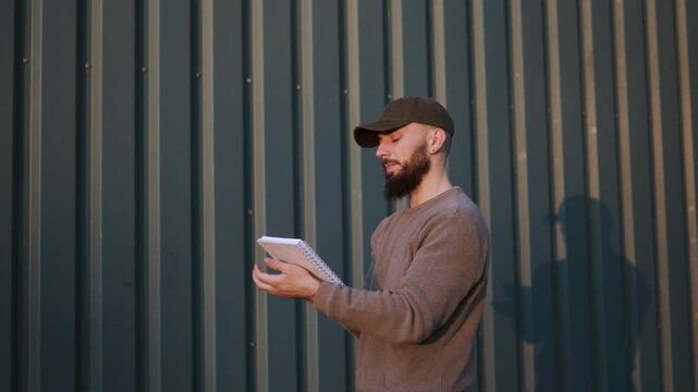 Creative bearded man in a cap thinking and writing notes in a notebook outdoors against a corrugated metal wall