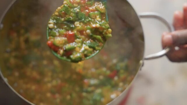 Black vendor ladling steaming vegetable stew, closeup of green ladle lifting chunky mix of corn, tomato, peppers and greens from metal pot hand holds handle, steam rising, market stall setting