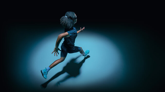 Female teenager athlete in dynamic running pose with futuristic visor in dark spotlight studio. Concept of performance analytics, modern technology, sport training, and digital fitness future.