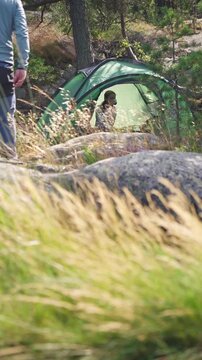 Couple in a green tent among rocks and pine trees in Finland, enjoying peaceful camping in the forest with tall grass in the foreground.