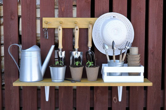 On the outside plank wall of firewood shed are hanging a shelf and a hanger with set of seedlings, pots, watering can, crate, old iron shears, keys and various gardening tools and supplies.