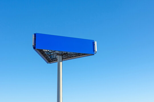 A blank blue triangular billboard stands tall against a clear, cloudless azure sky, featuring integrated telecommunications antennas on its sleek modern structure.