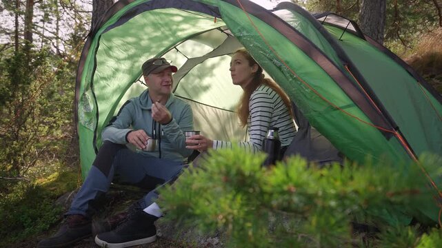 Adult couple talking and drinking tea inside a tent with a scenic coastal view. Authentic camping lifestyle, connection, and slow travel adventure during summer vacation.