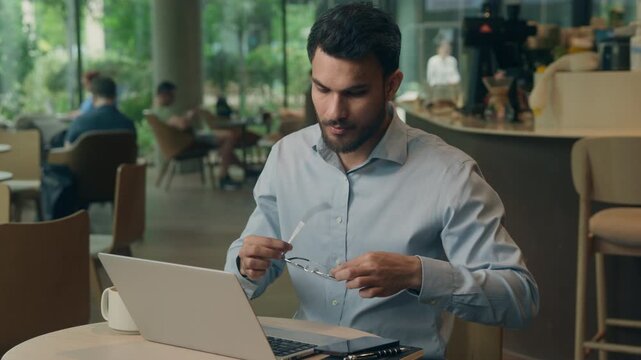 Sleepy napping tired Indian business man at table with laptop putting on glasses starting working computer bored businessman Arabian Indian male guy feeling chronic fatigue insomnia in cafe workplace