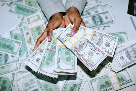 Black woman holding stacks of hundred dollar bills with both hands, displaying large amount of cash on table covered with bundles of United States currency, close up of hands