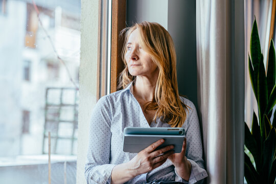 Smiling caucasian woman in her 50s holding tablet at home near window