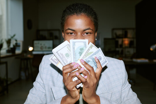 Portrait of young adult Black woman smiling and holding several stacks of US dollar bills in both hands, standing in modern office environment with computer monitors visible in background