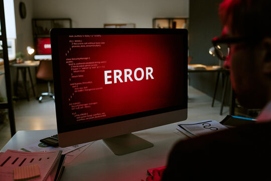 Caucasian middle aged man sitting at desk facing computer monitor displaying large error message on red screen, working in modern office environment with documents and stationery visible