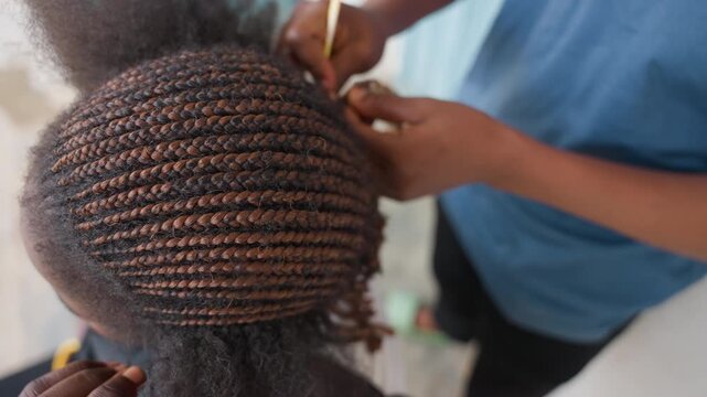 Black stylist braiding neat cornrows overhead closeup of skilled hands forming precise rows on textured hair, focused studio atmosphere, natural light highlighting brown tones and scalp detail