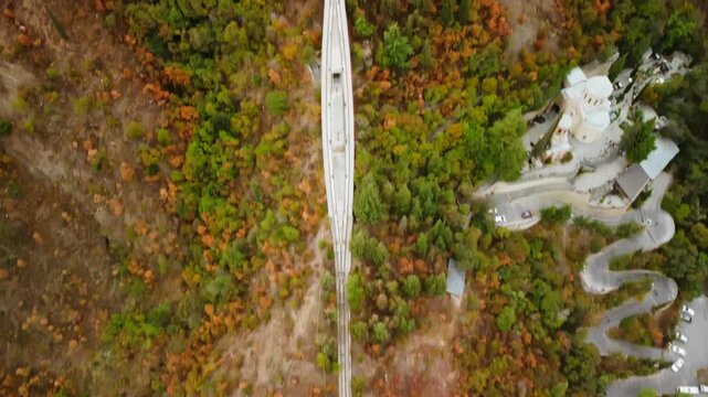 Aerial bridge over dense autumn fores leading to old building on the hill next to tele tower filmed by drone. Landscape of Tbilisi nature green and orange foliage and rocky tower. Travel to Georgia