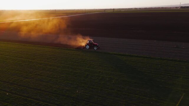 Aerial view of a red tractor tilling a vast field, stirring up dust clouds against the backdrop of a sunset in Jazak, Vojvodina, Serbia.