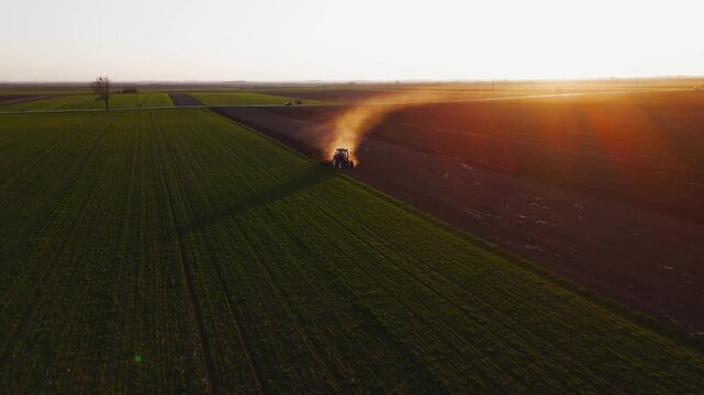 Aerial view of a tractor tilling the soil, creating a cloud of dust, amidst the green fields at sunset, Jazak, Vojvodina, Serbia.