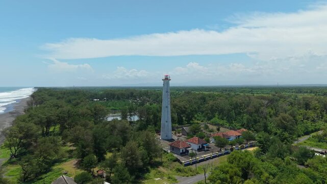 Aerial view of Mercusuar Samas Lighthouse, a tall white structure rising above lush green trees near the dark sandy beach, Bantul Regency, Daerah Istimewa Yogyakarta, Indonesia.