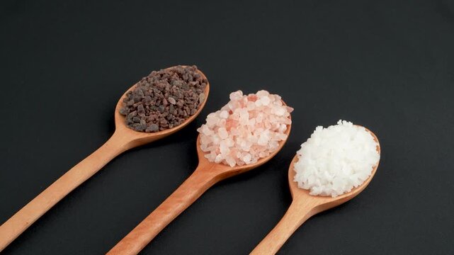 Close up diagonal rotating macro of three gourmet coarse salts (black, pink, white) on rustic wooden spoons isolated on black. Premium food texture.