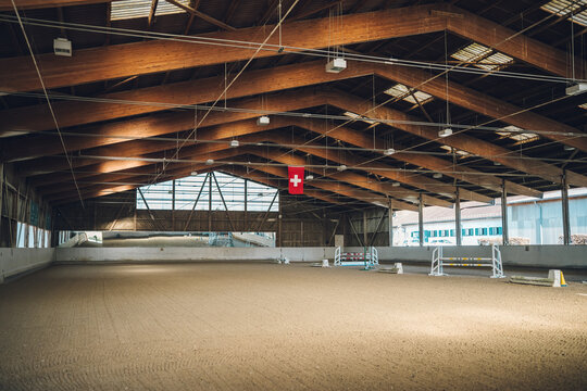 Riding arena featuring sand footing, wooden beam ceiling, and equestrian hurdles, with a swiss flag hanging