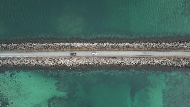 Aerial view of the bridge at Flakstadbruene, where the turquoise waters meet the rocky causeway road with a car driving on it, Flakstadbruene, Nordland, Norway.