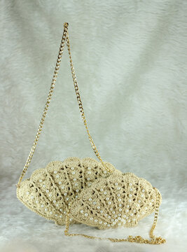 Beige crocheted shoulder bag in the shape of a seashell with a gold-colored handle sits on a white fluffy blanket. The bag features pearl bead details.
