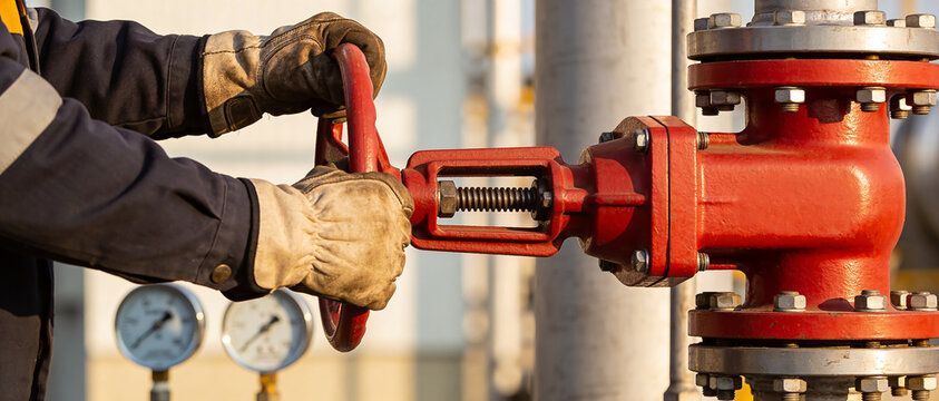 Worker's hands in protective gloves turning red valve handwheel on industrial pipeline.