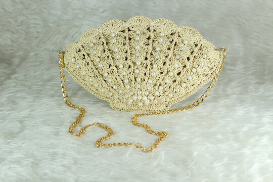Beige crocheted shoulder bag in the shape of a seashell with a gold-colored handle sits on a white fluffy blanket. The bag features pearl bead details.	
