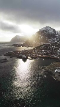 Aerial view of the rugged coastline meeting the tranquil sea, with sunlight piercing through clouds over Tind, Nordland, Norway.