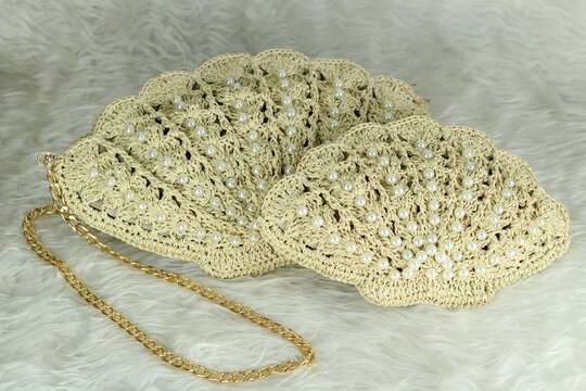 Beige crocheted shoulder bag in the shape of a seashell with a gold-colored handle sits on a white fluffy blanket. The bag features pearl bead details.	